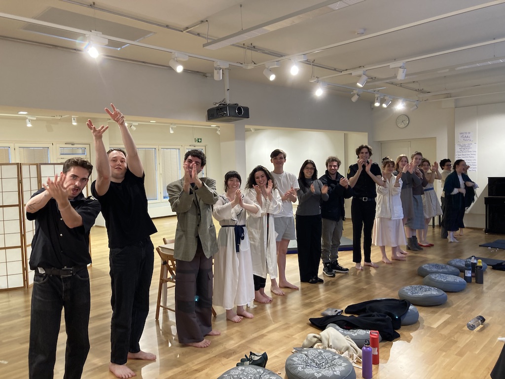 Candid photo of the spring 2024 Stanislavski, Brecht, and Beyond class. Students are in a bright rehearsal studio, standing in a curtain call line, smiling and applauding at the end of their presentation of scenes from Ödön von Horváth’s 'Faith Hope Charity'. Cushions, shoes, and water bottles are scattered on the floor in front of the students.